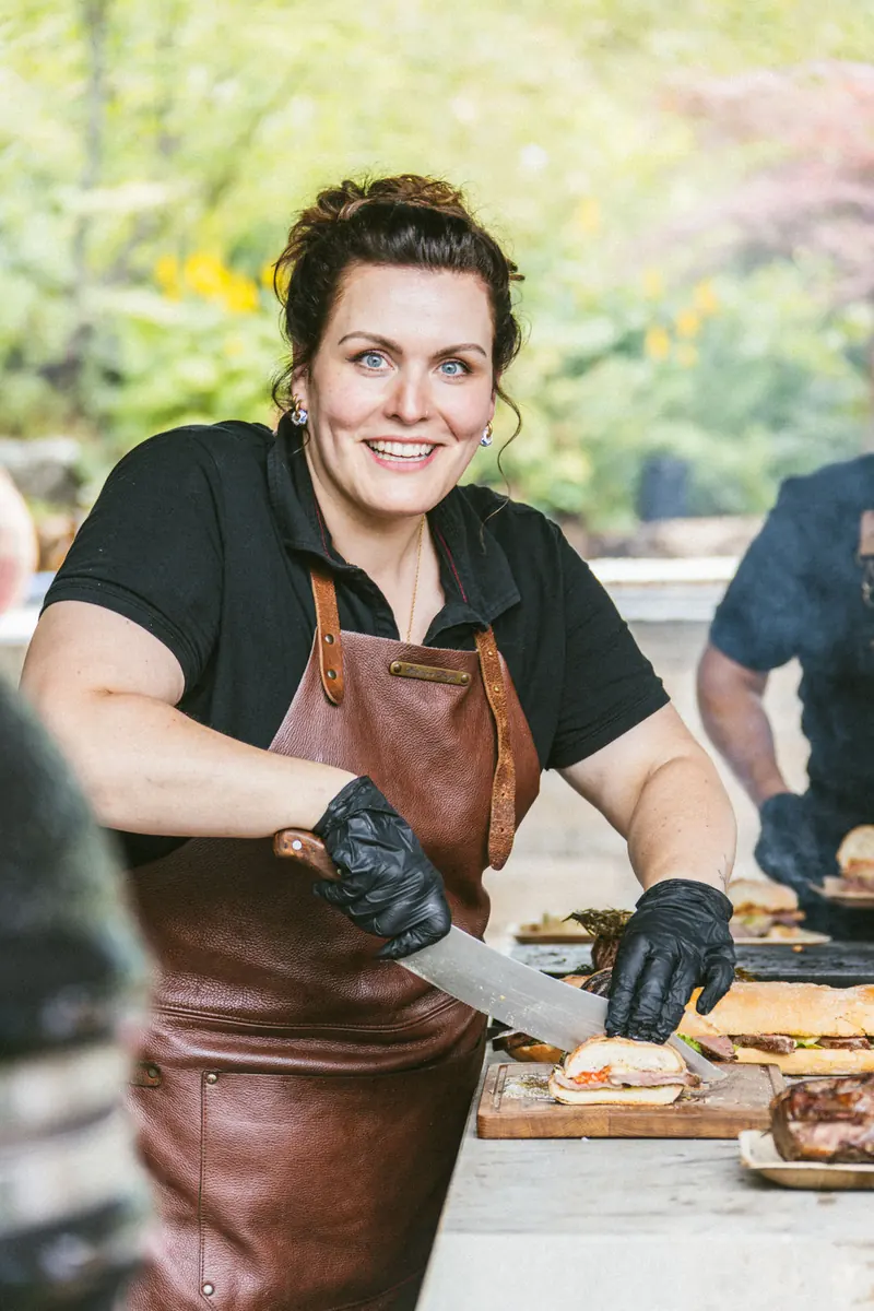 Katharina Bertl in Lederschürze am Grill, sie schneidet Fleisch auf einem Holzbrett