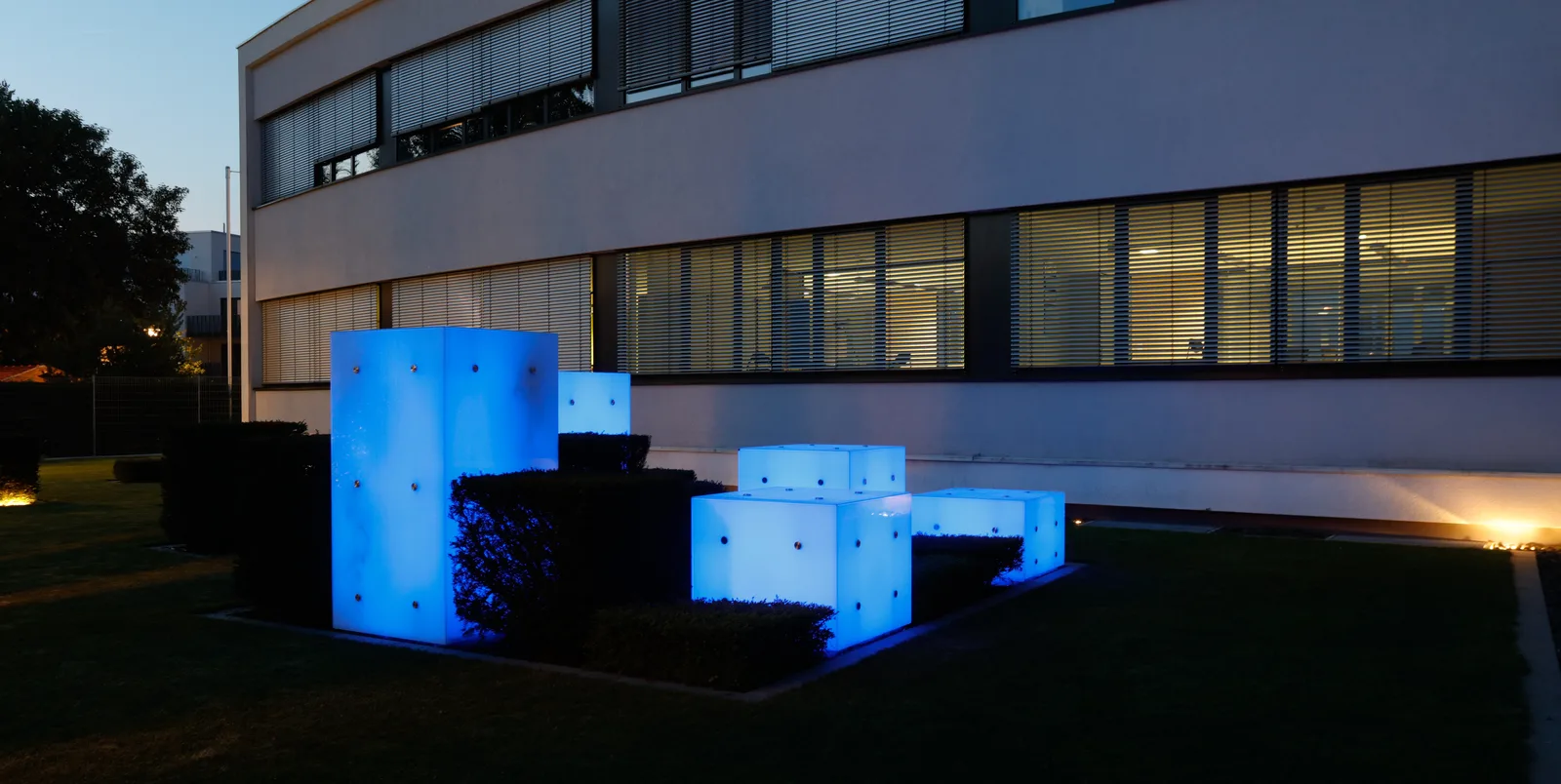 Sitzwürfel, die selbst leuchten: blau-weiße Lichtkuben vor der Fassade des Bürogebäudes