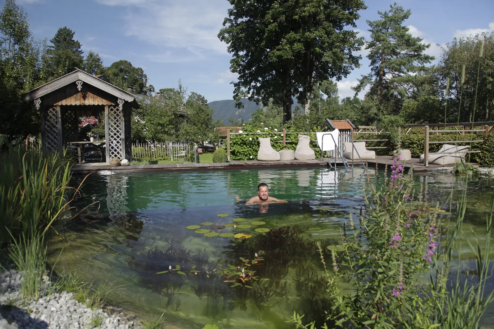 Fred Fuchs schwimmt im Schwimmteich am Landhaus Benediktenhof, dahinter Liegen, Badehaus und die Berge