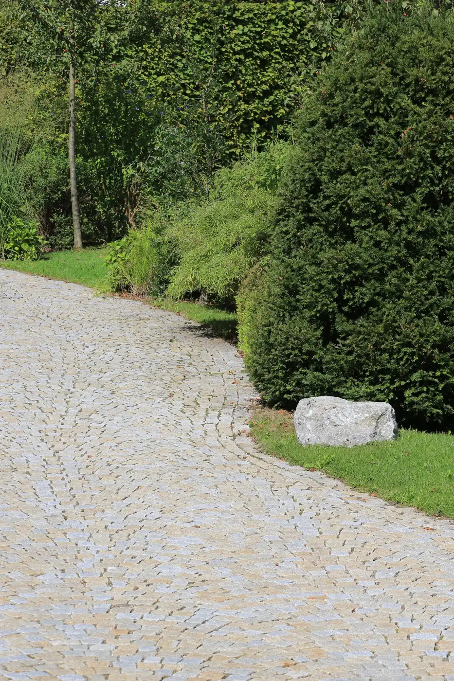 Der gepflasterte Weg schwingt in Bögen an den Eiben vorbei, ein Findling markiert die Kante