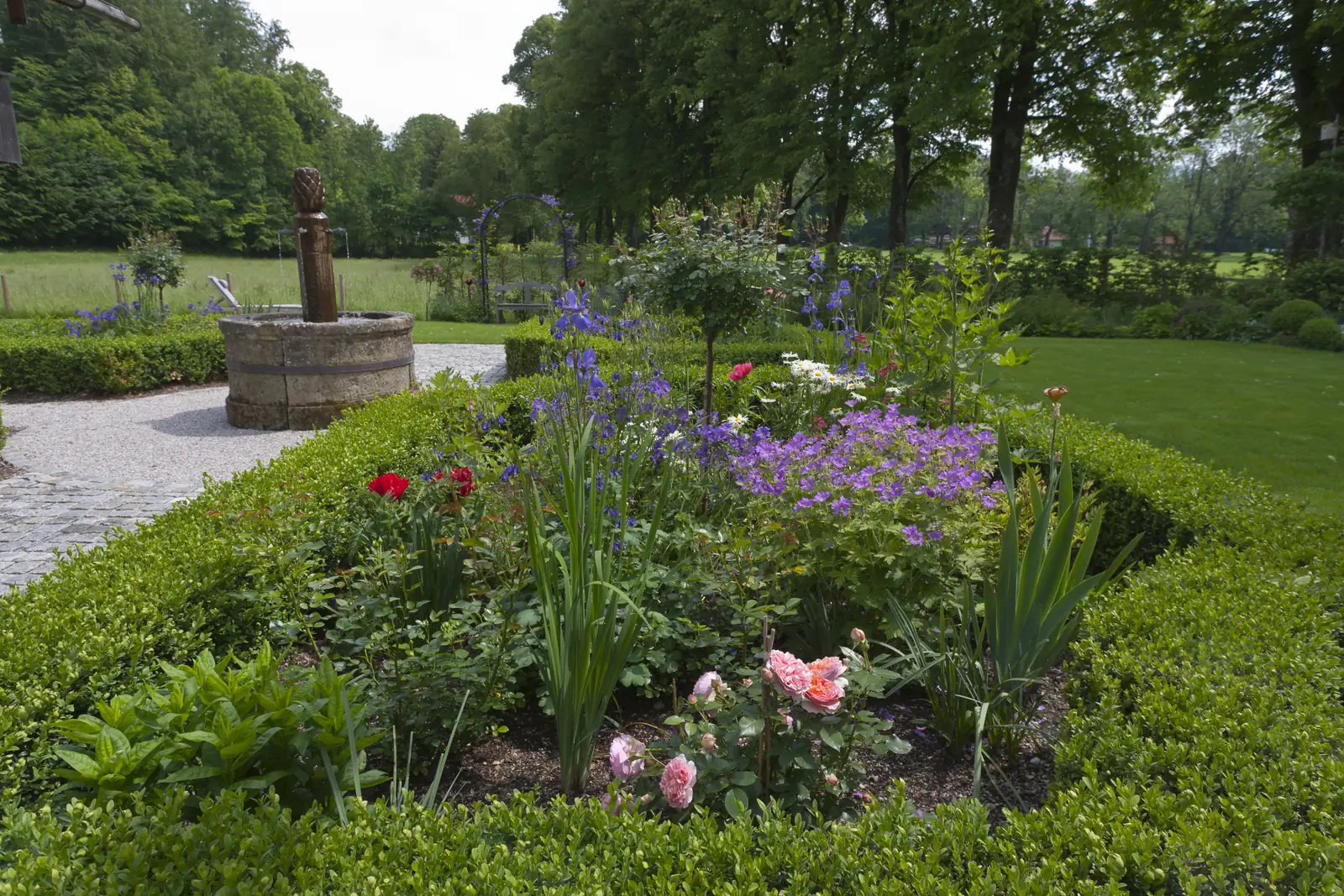 Der Brunnen aus Naturstein steht im Staudenbeet, Rosen blühen davor