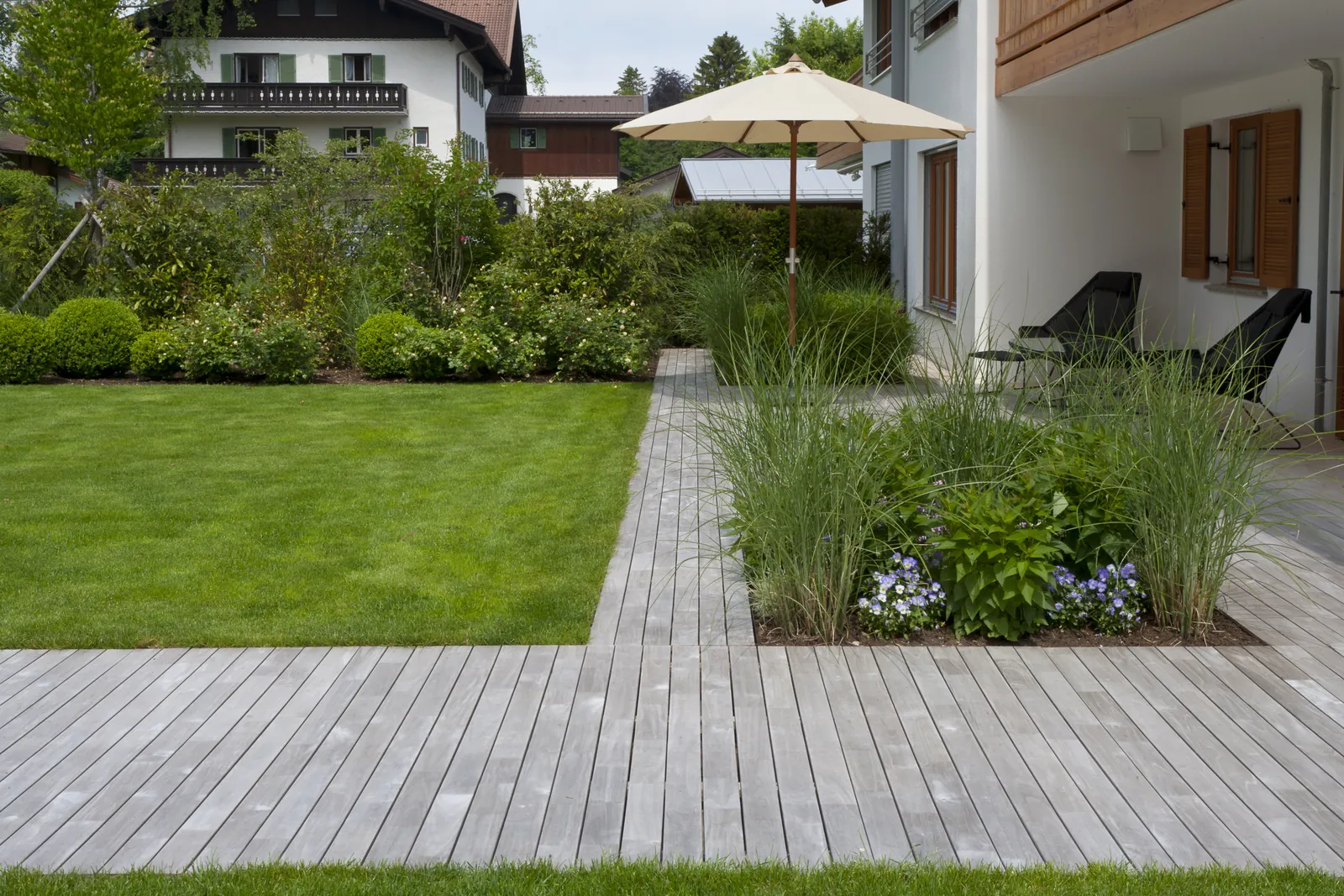 Holzdeck und Rasen verzahnen sich, dazwischen Gräser und Pflanzinseln
