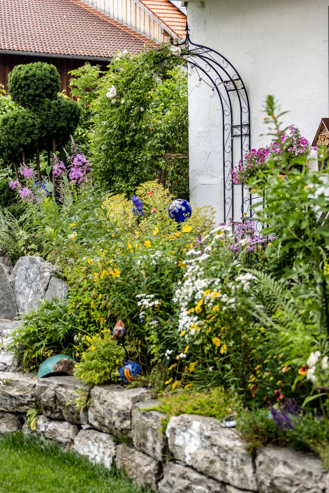 Stauden in voller Blüte quellen über die Mauerkrone, dazwischen blaue Glaskugeln