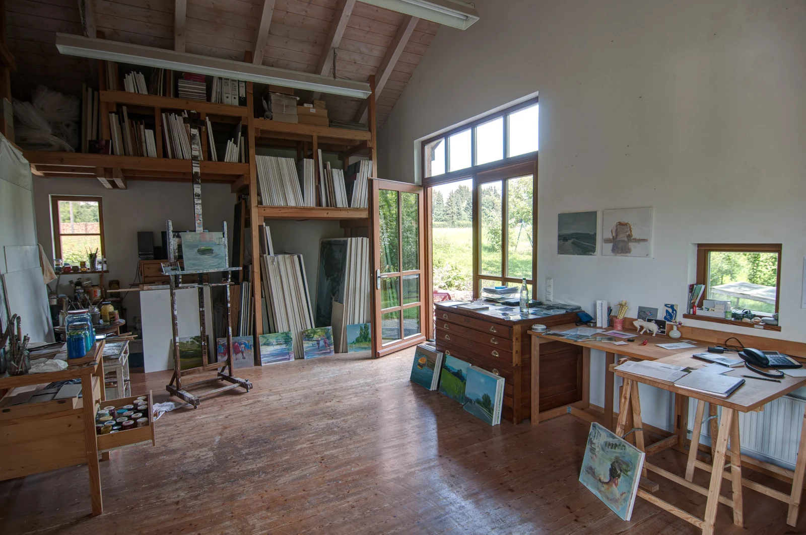 Das Atelier: Staffeleien, Bilder an der Wand und die Fensterfront zum Garten