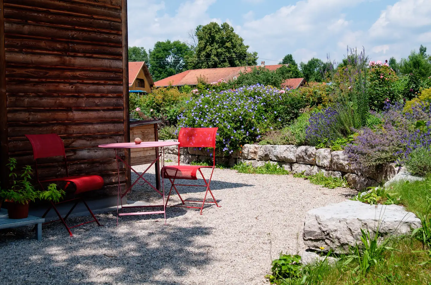 Sitzplatz mit roten Metallmöbeln an der Holzwand, ringsum Stauden im Kies