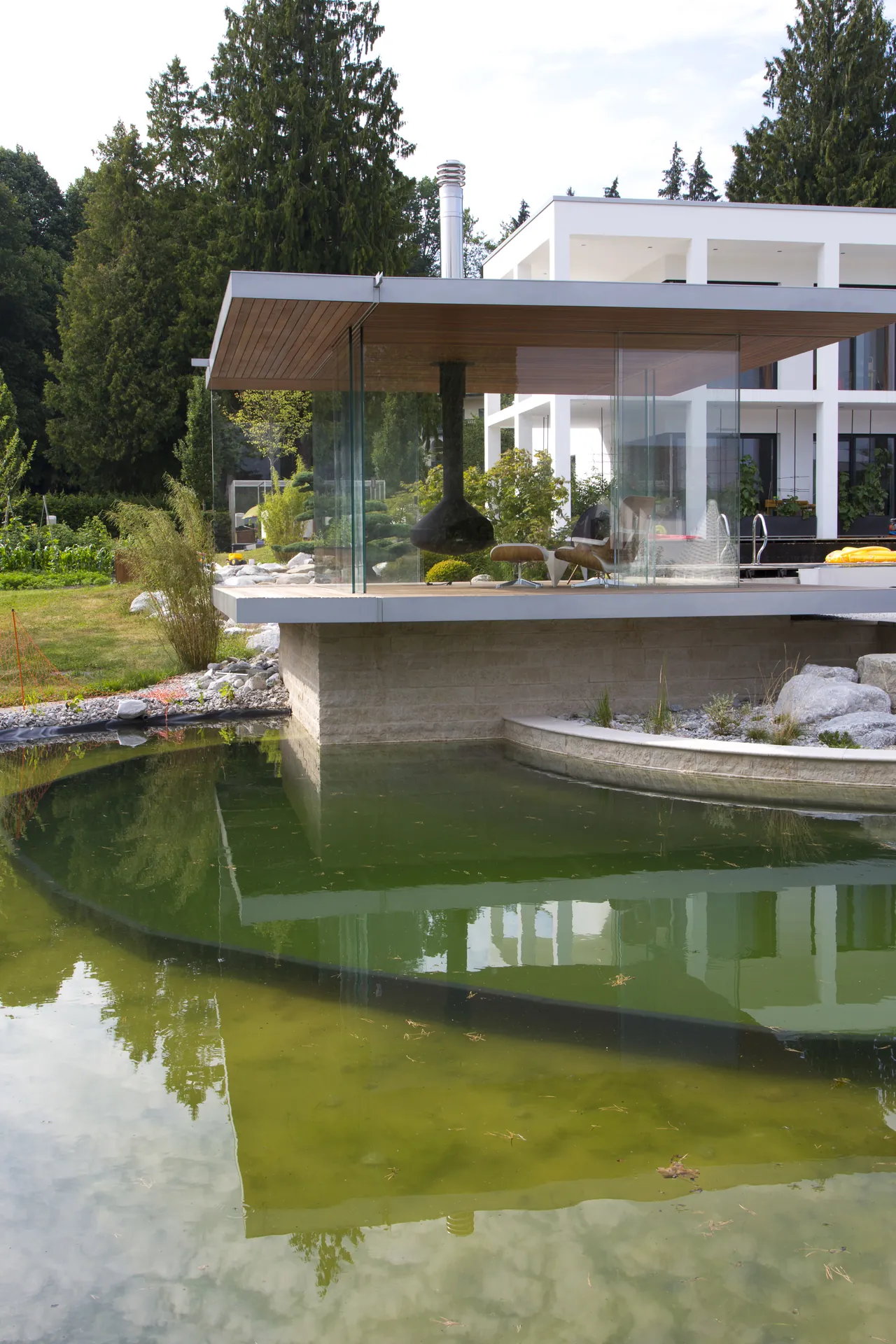 Die auskragende Sichtbetonplatte über dem Wasser, im Teich spiegeln sich Pavillon und Bäume