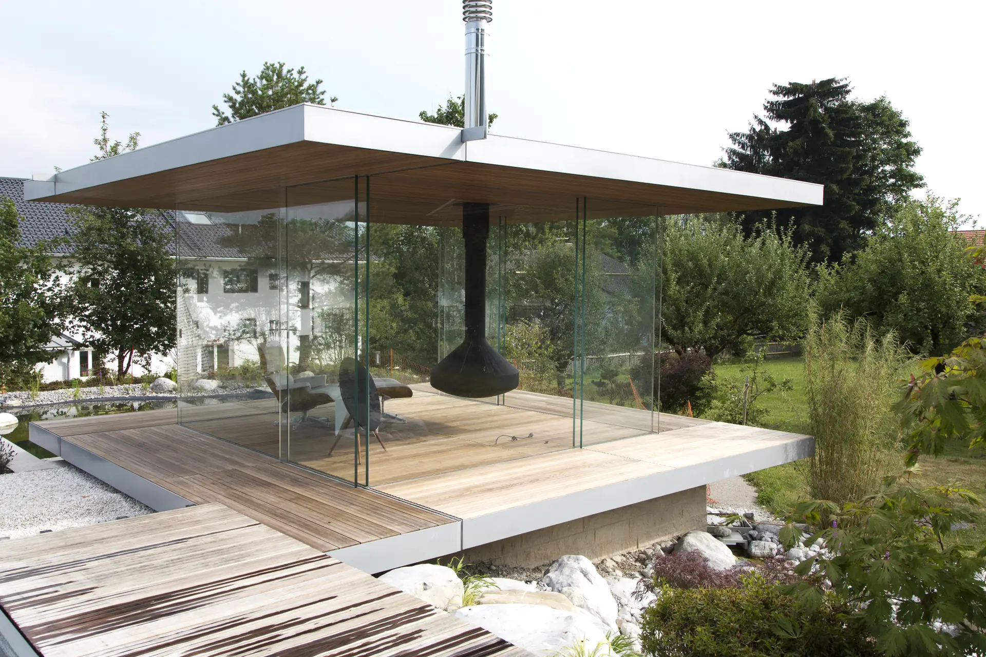 Der gläserne Pavillon auf dem Holzdeck, darin der schwarze abgehängte Kamin