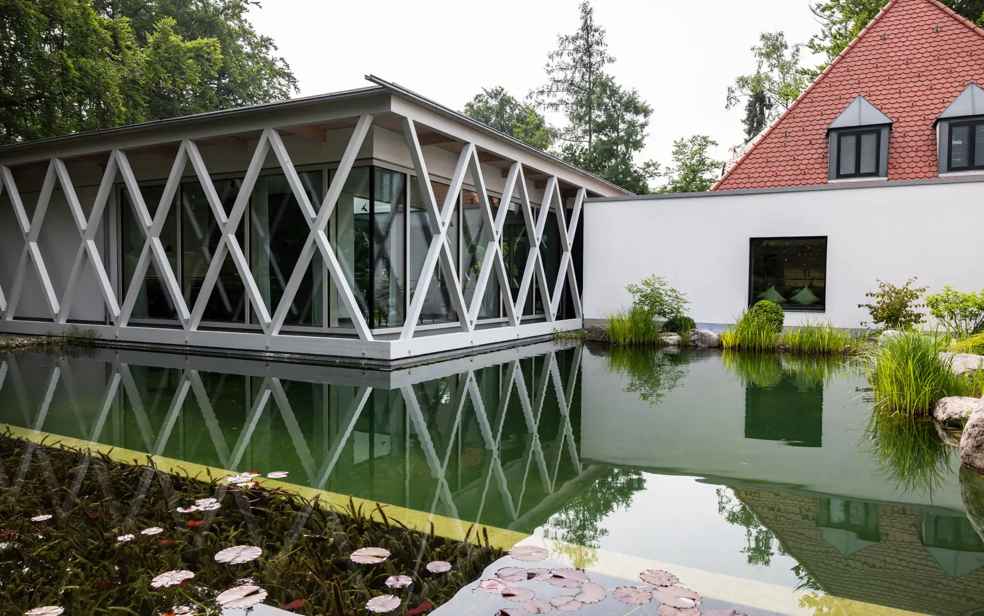 Die weiße Rautenfassade spiegelt sich im Schwimmteich, am Ufer Seerosenblätter und Schilf