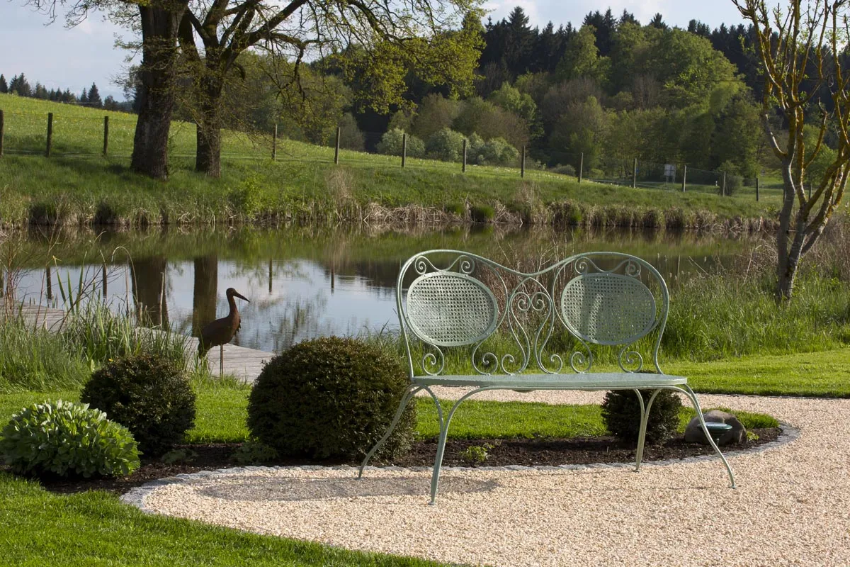 Die Bank am Kiesplatz über dem Wasser, daneben ein Storch aus Metall
