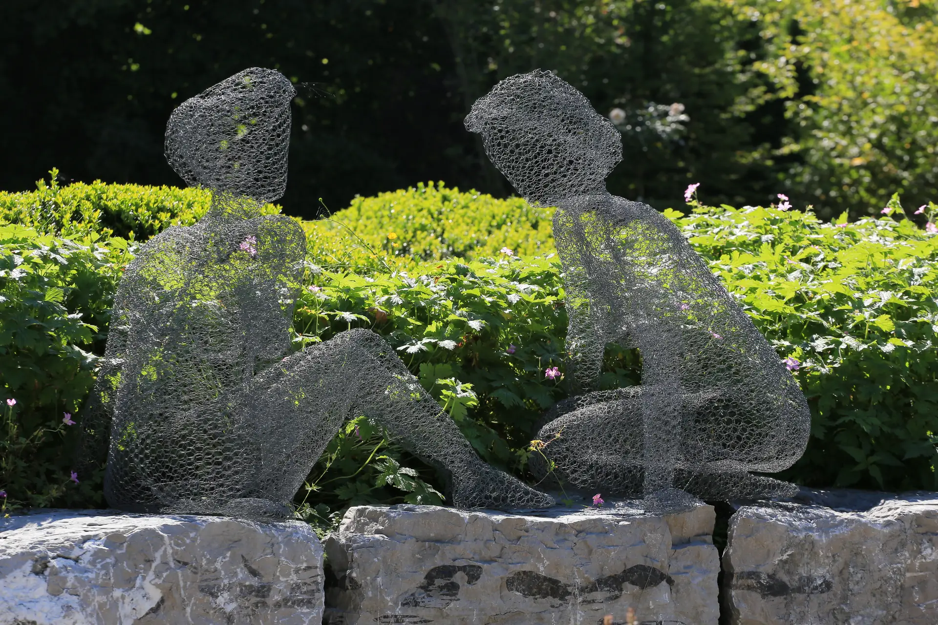 Zwei sitzende Figuren aus Drahtgeflecht auf der Natursteinmauer