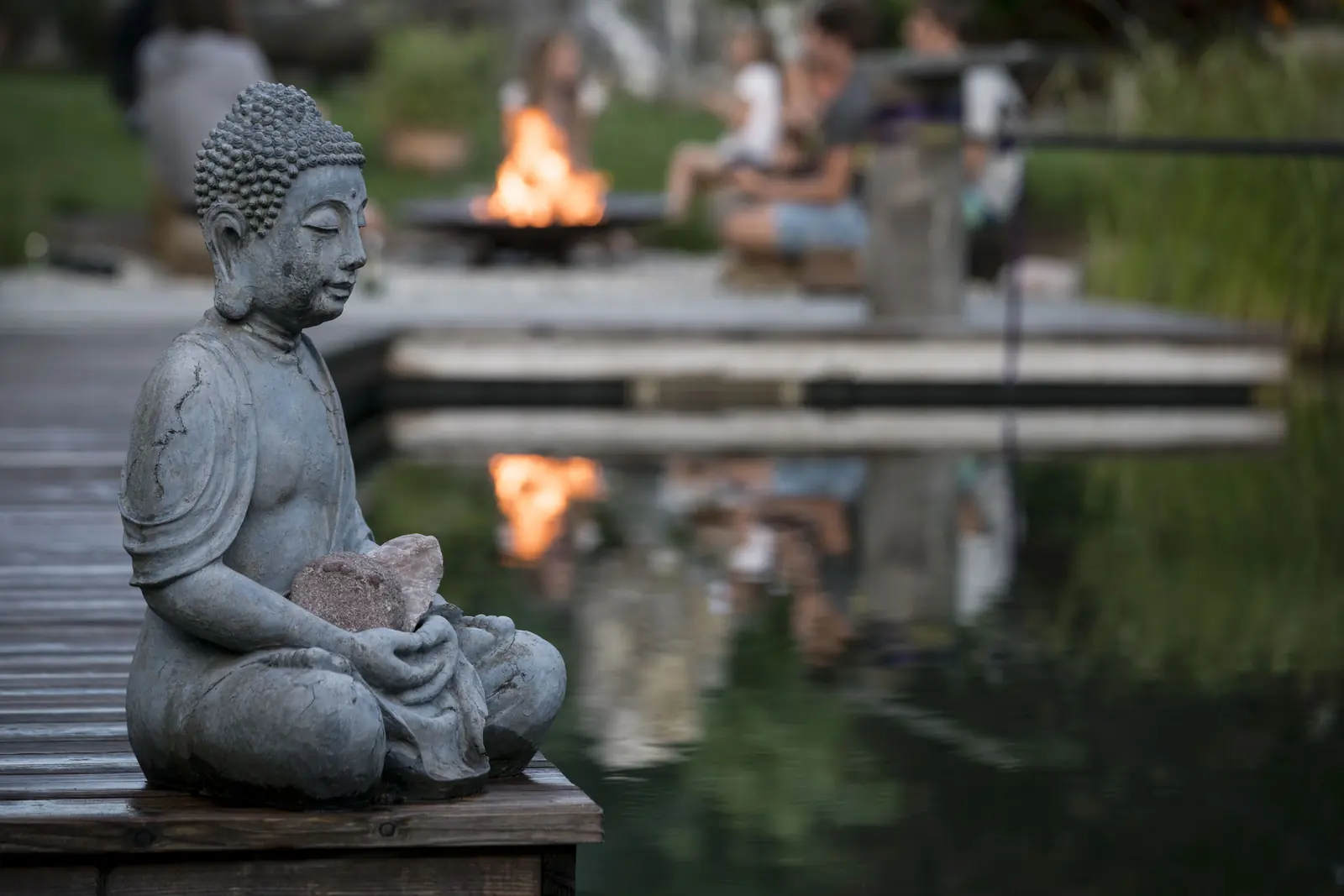 Eine Buddha-Figur am Holzsteg, dahinter unscharf das Feuer in der Schale