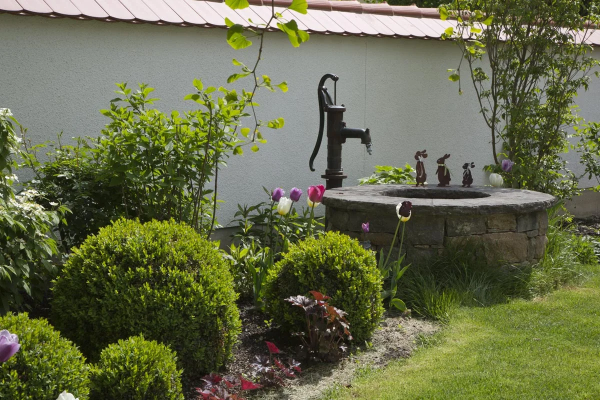 Eine alte Handschwengelpumpe auf einem runden Granitbrunnen zwischen Formschnittkugeln