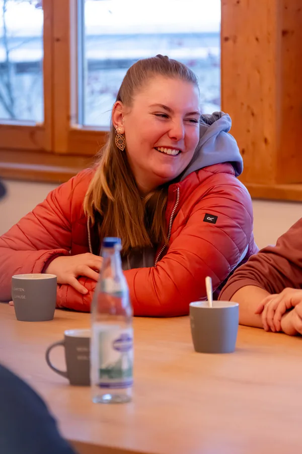 Kollegin lacht am Tisch, Kaffee und Wasserflasche vor sich