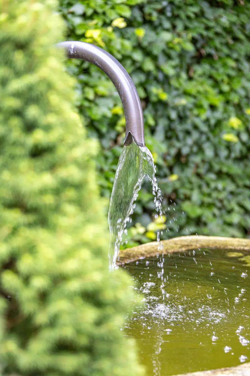 Wasser läuft aus einem dunklen Auslass in einen Natursteintrog, dahinter eine grüne Wand